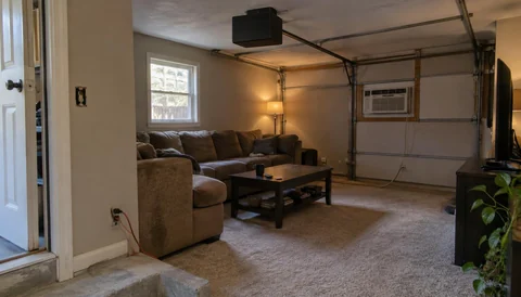 Garage converted into a carpeted living room, with the old garage door tracks and a window air conditioner still in place