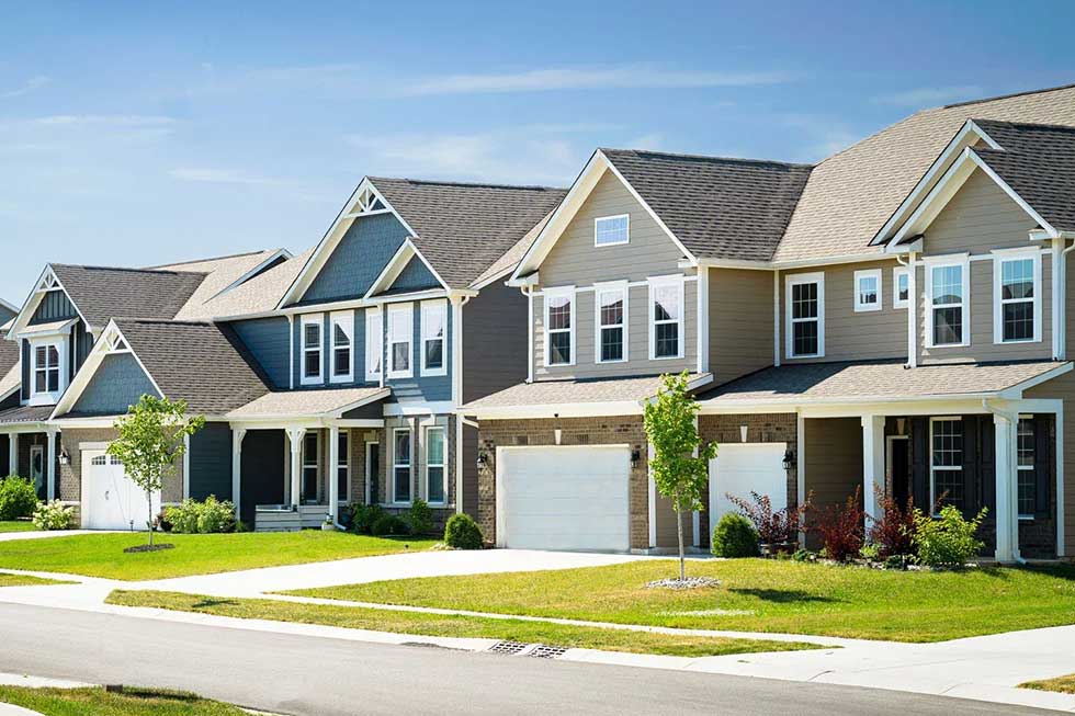 A row of modern suburban houses with garages, well-maintained lawns, trees, and a clear blue sky overhead